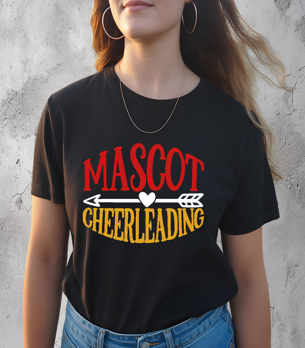 Woman wearing black t-shirt with bold red and yellow “Mascot Cheerleading” design and heart-arrow graphic, standing against a light textured wall background.