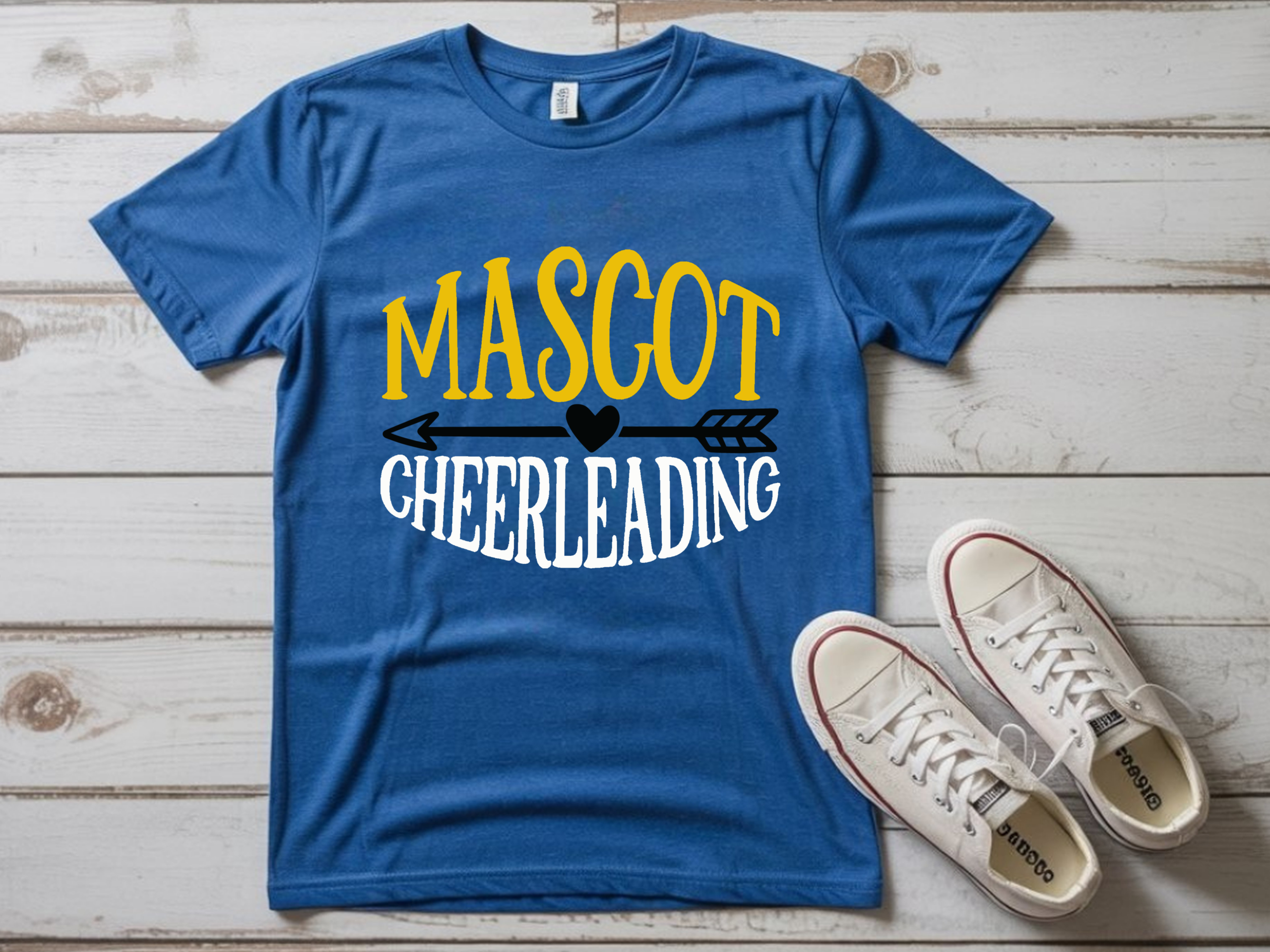 Blue t-shirt with bold yellow and white “Mascot Cheerleading” design featuring arrow and heart graphic, displayed flat with white sneakers on wooden background.