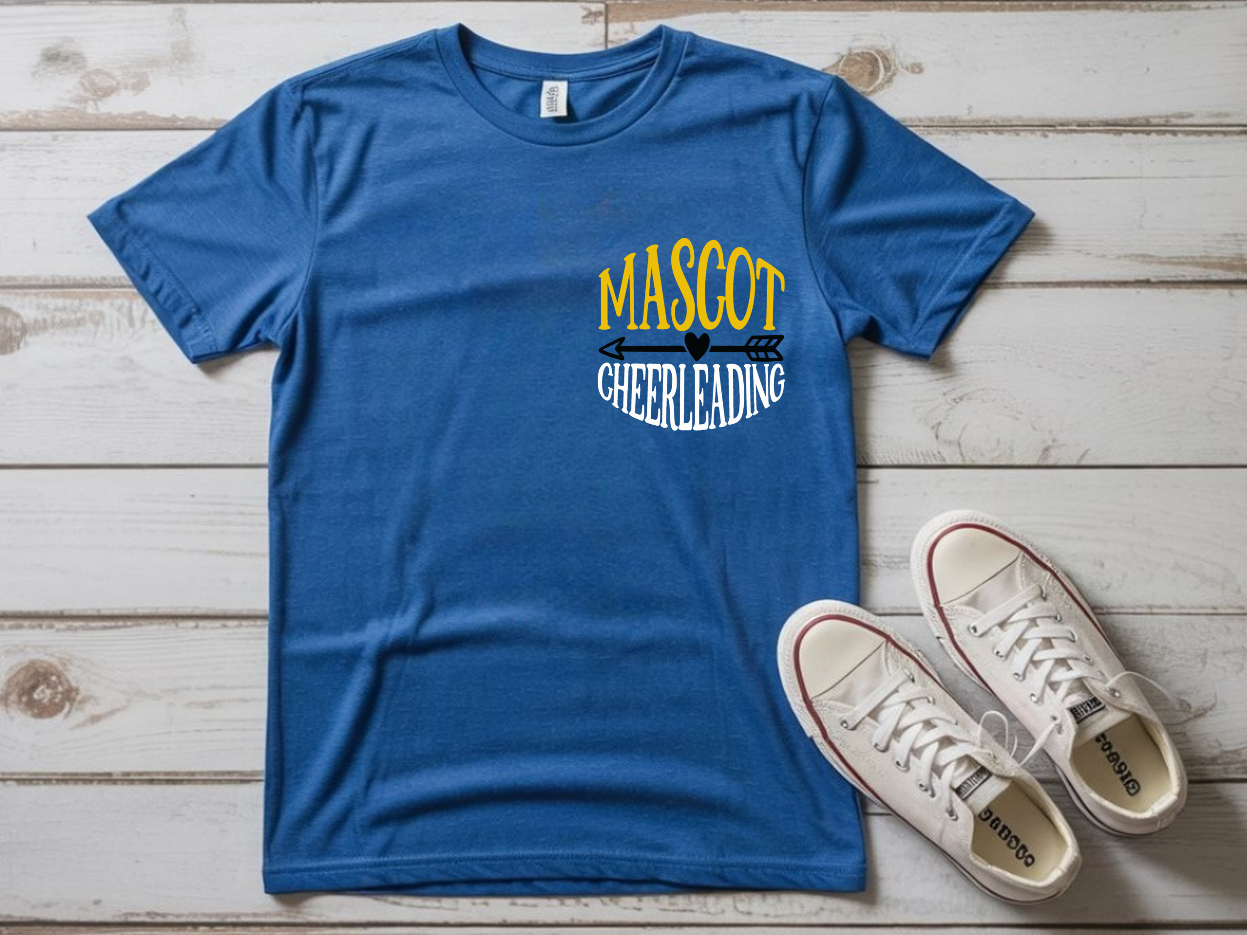 Blue t-shirt with bold yellow and white “Mascot Cheerleading” design featuring arrow and heart graphic on the front left side pocket of the shirt, displayed flat with white sneakers on wooden background.