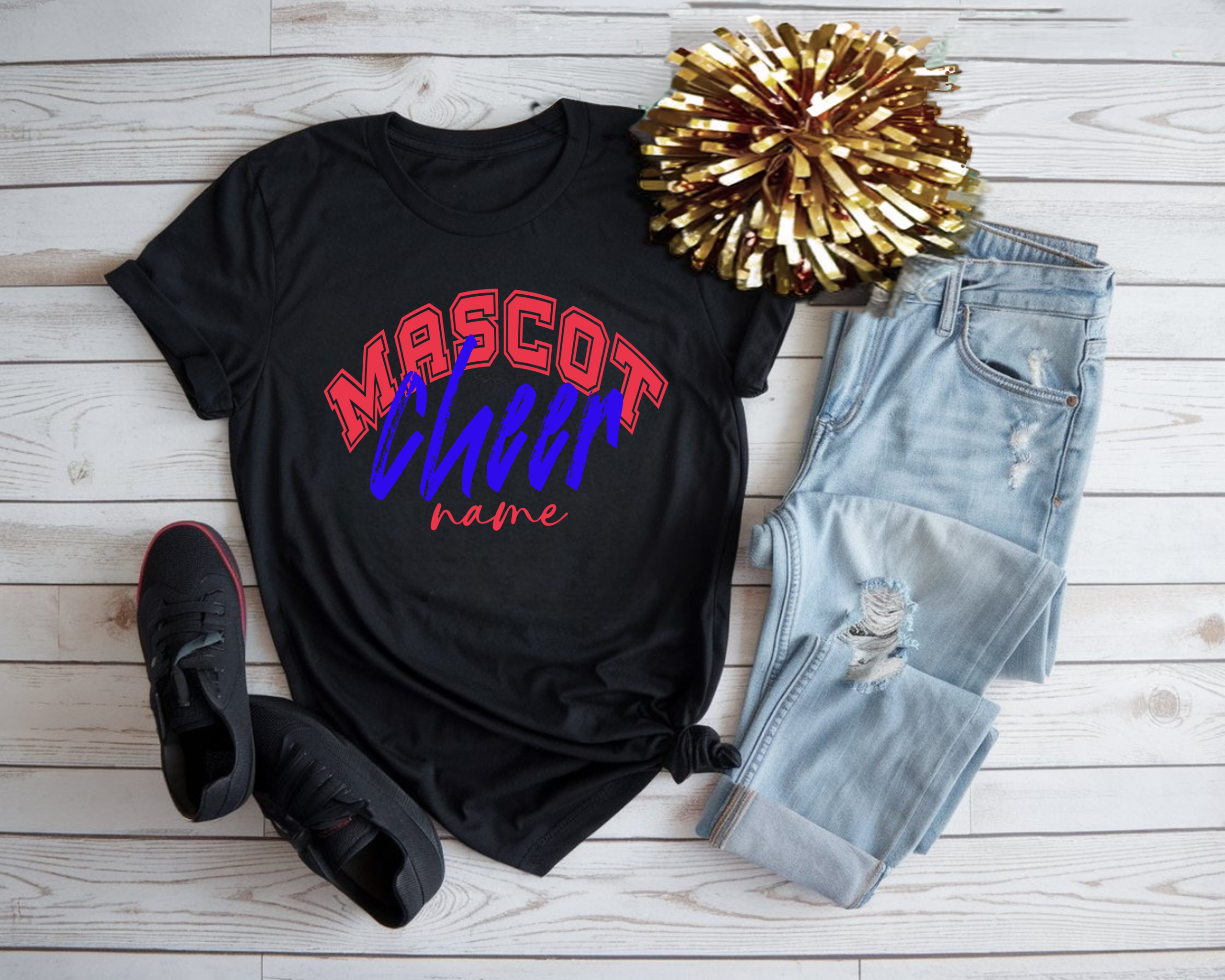 Black custom cheerleading t-shirt with red, blue, and white “Mascot Cheer Name” design, styled with jeans, black sneakers, and a gold pom-pom on a white wood background.