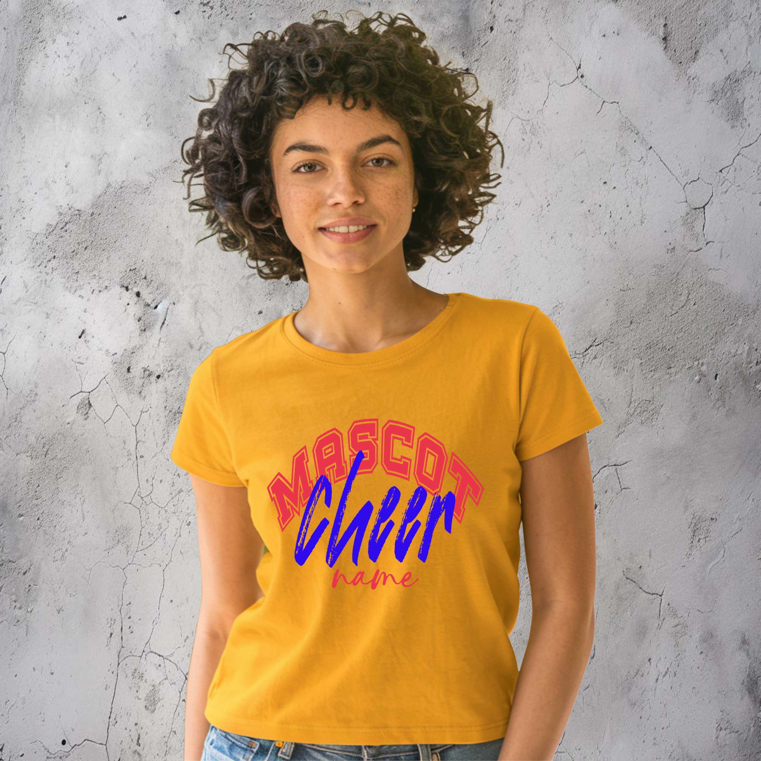 Smiling woman wearing a mustard yellow custom cheer shirt with red, blue, and pink “Mascot Cheer Name” design, standing against a light gray textured wall.