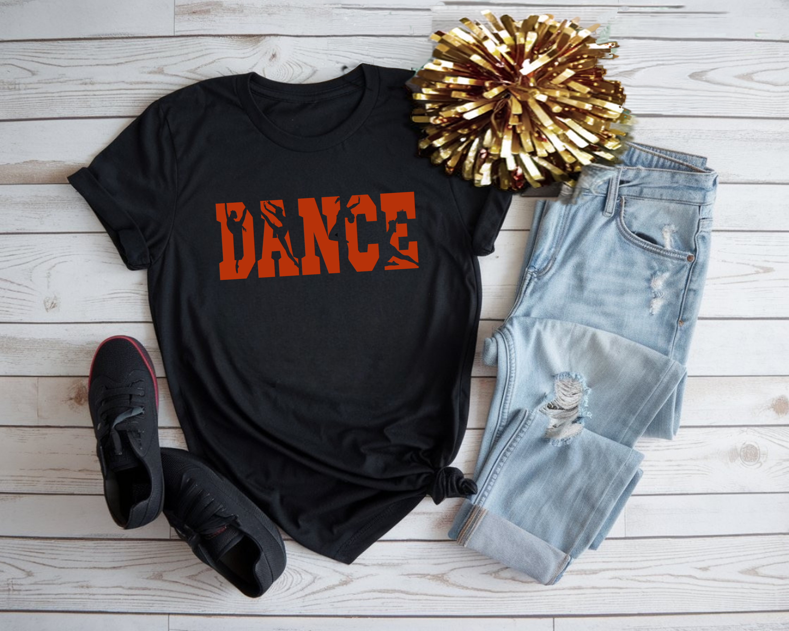 Black dance t-shirt with bold orange varsity “DANCE” lettering featuring dancer silhouettes, styled flat lay with light ripped jeans, black sneakers, and gold pom pom on a white wood background.