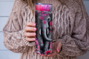 A skinny tumbler with a detailed floral elephant design in pink and gray tones, held in someone’s hands, choose to add text and placement of text.