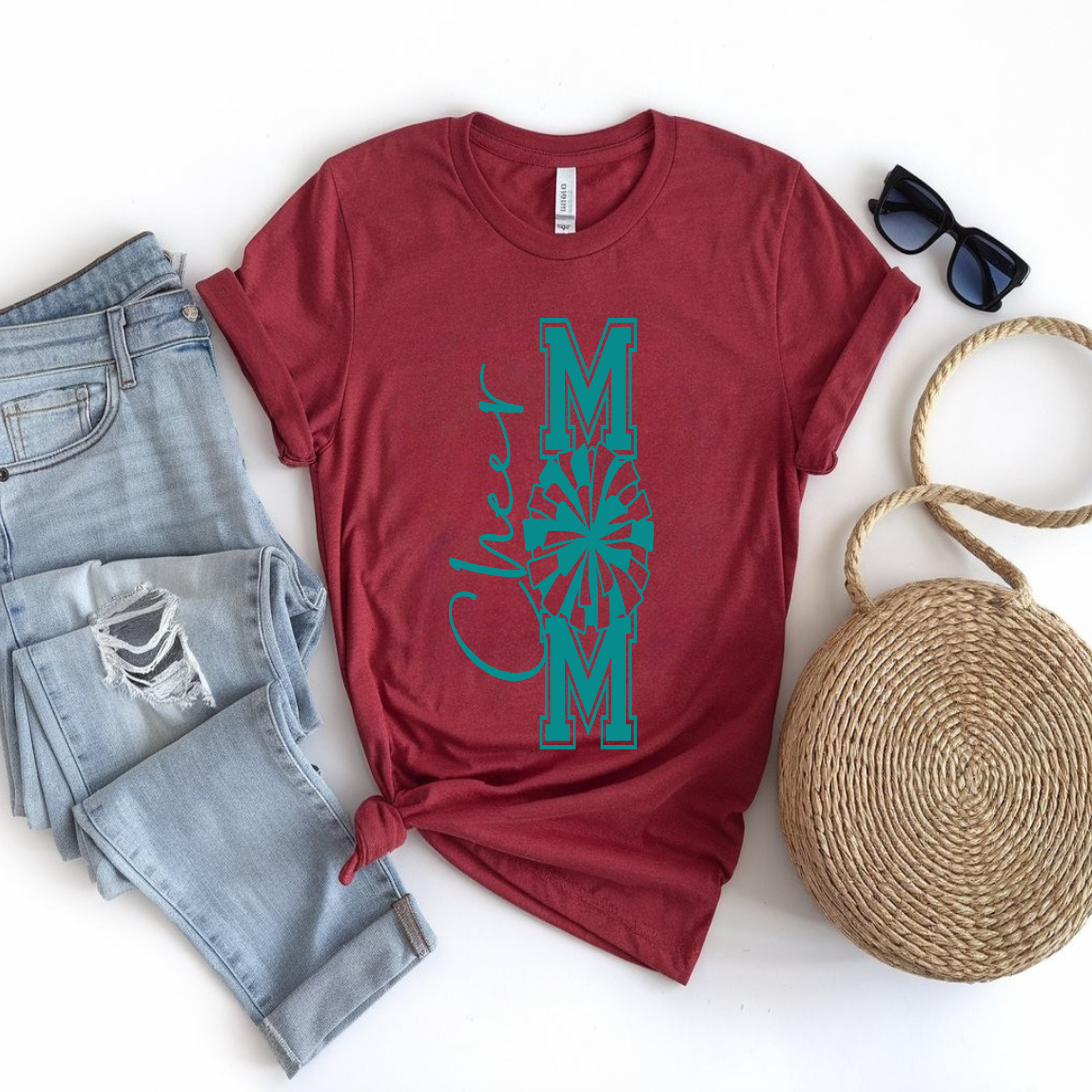 Laying flat on the floor is a red “Cheer Mom” t‑shirt with teal lettering and a pom‑pom graphic, styled with ripped light blue jeans, black sunglasses, and a round woven bag on a white background.