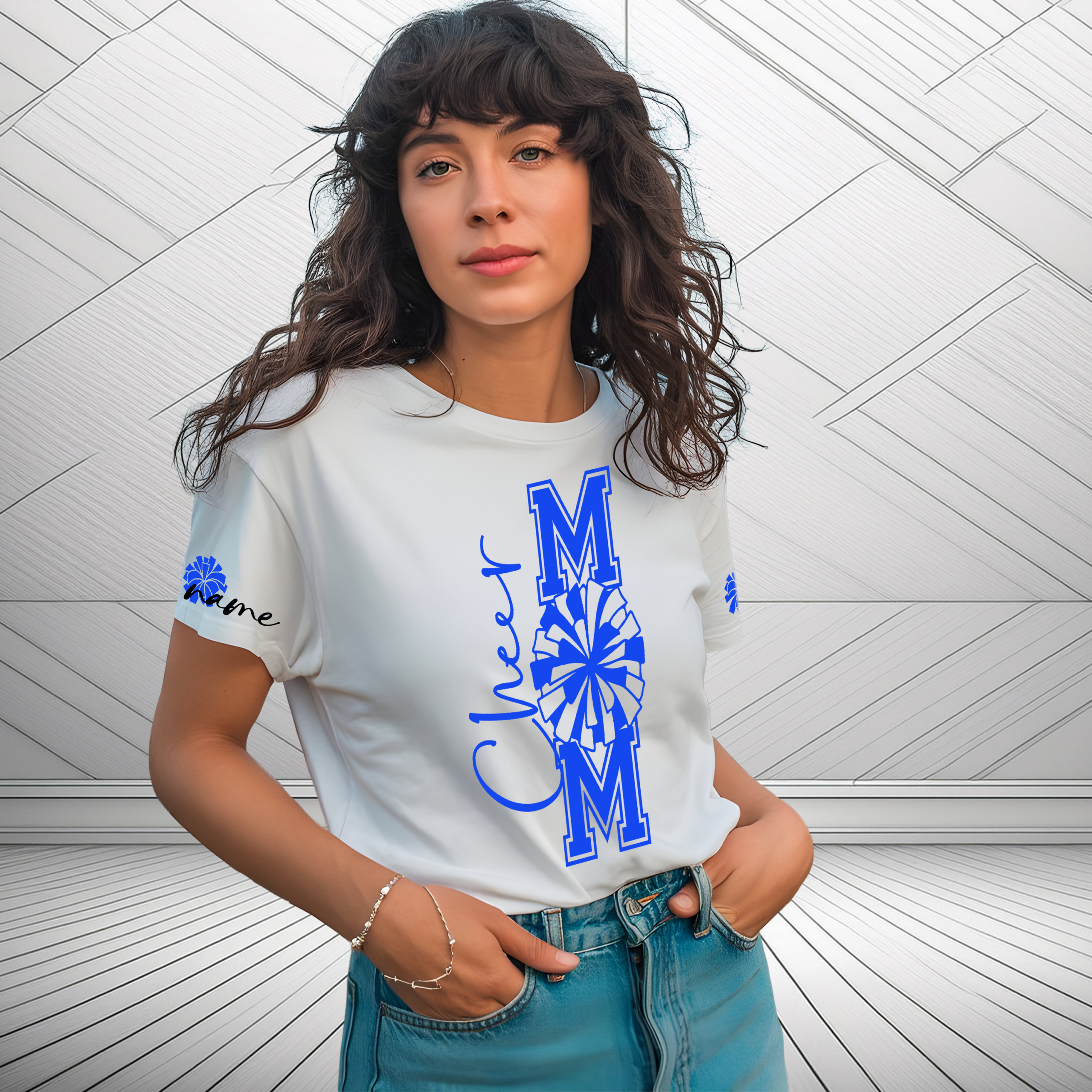Woman wearing a white custom “Cheer Mom” t‑shirt with blue varsity lettering, pom‑pom design, and personalized name on the sleeve, standing against a light geometric wall background.