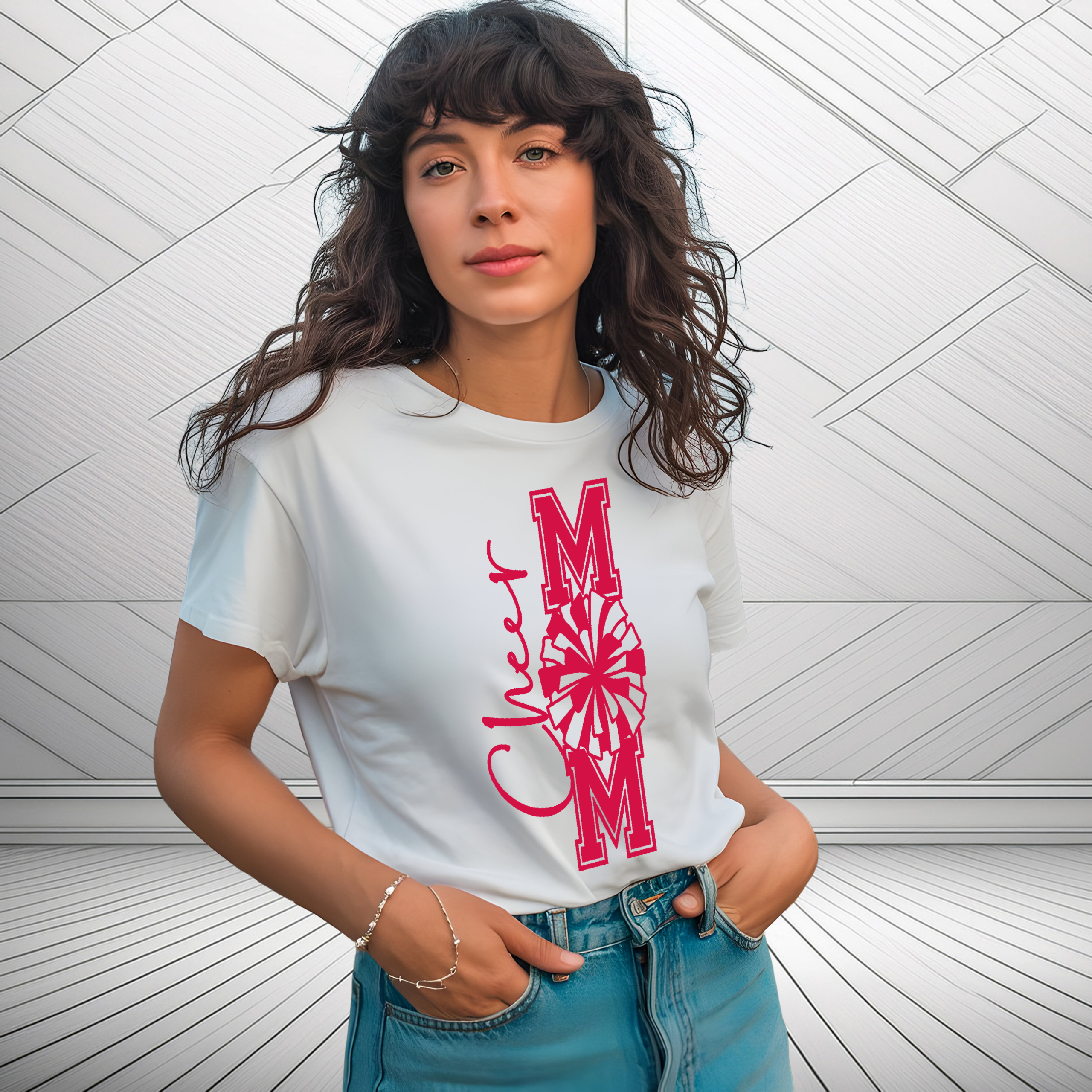Woman wearing a white “Cheer Mom” t‑shirt with red varsity lettering and pom‑pom graphic, standing in front of a modern gray geometric wall while posing confidently with one hand in pocket.