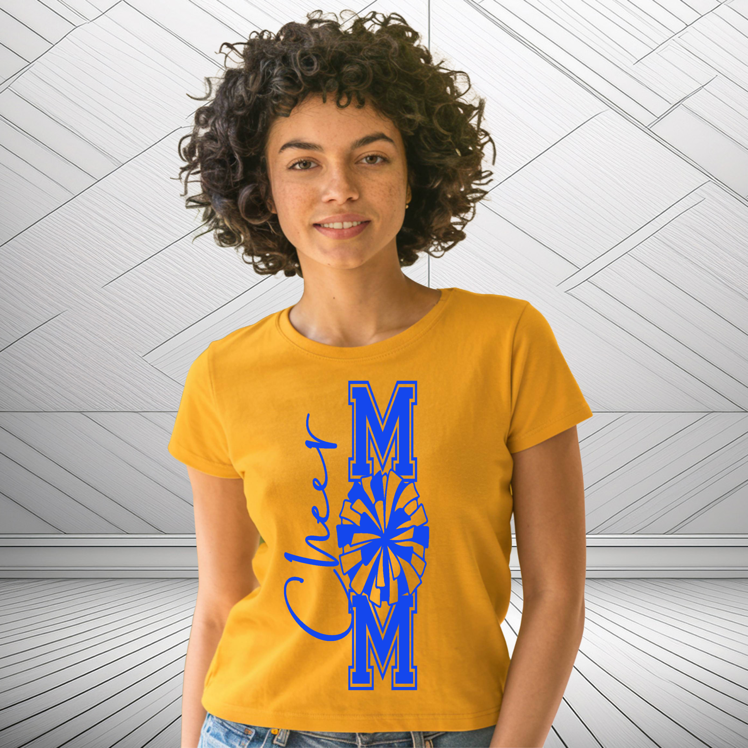 Woman wearing a gold “Cheer Mom” t‑shirt with bold blue varsity lettering and pom‑pom design, standing in front of a modern white geometric wall background.