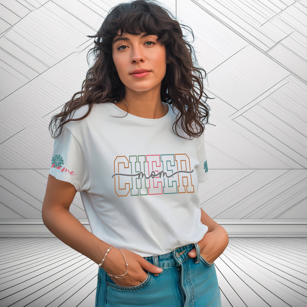Woman wearing a white “Cheer Mom” t‑shirt with bold multicolor varsity lettering and personalized name detail on the sleeves, standing against a pom.