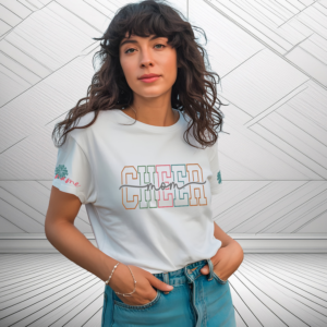 Woman wearing a white “Cheer Mom” t‑shirt with bold multicolor varsity lettering and personalized name detail on the sleeves, standing against a pom.