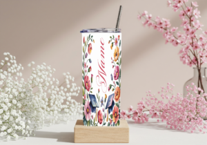 Custom personalized graduation skinny tumbler with watercolor floral design, graduation caps, and cursive name in pink on a white background, displayed with a metal straw on a wooden stand surrounded by pink and white flowers