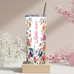 Custom personalized graduation skinny tumbler with watercolor floral design, graduation caps, and cursive name in pink on a white background, displayed with a metal straw on a wooden stand surrounded by pink and white flowers
