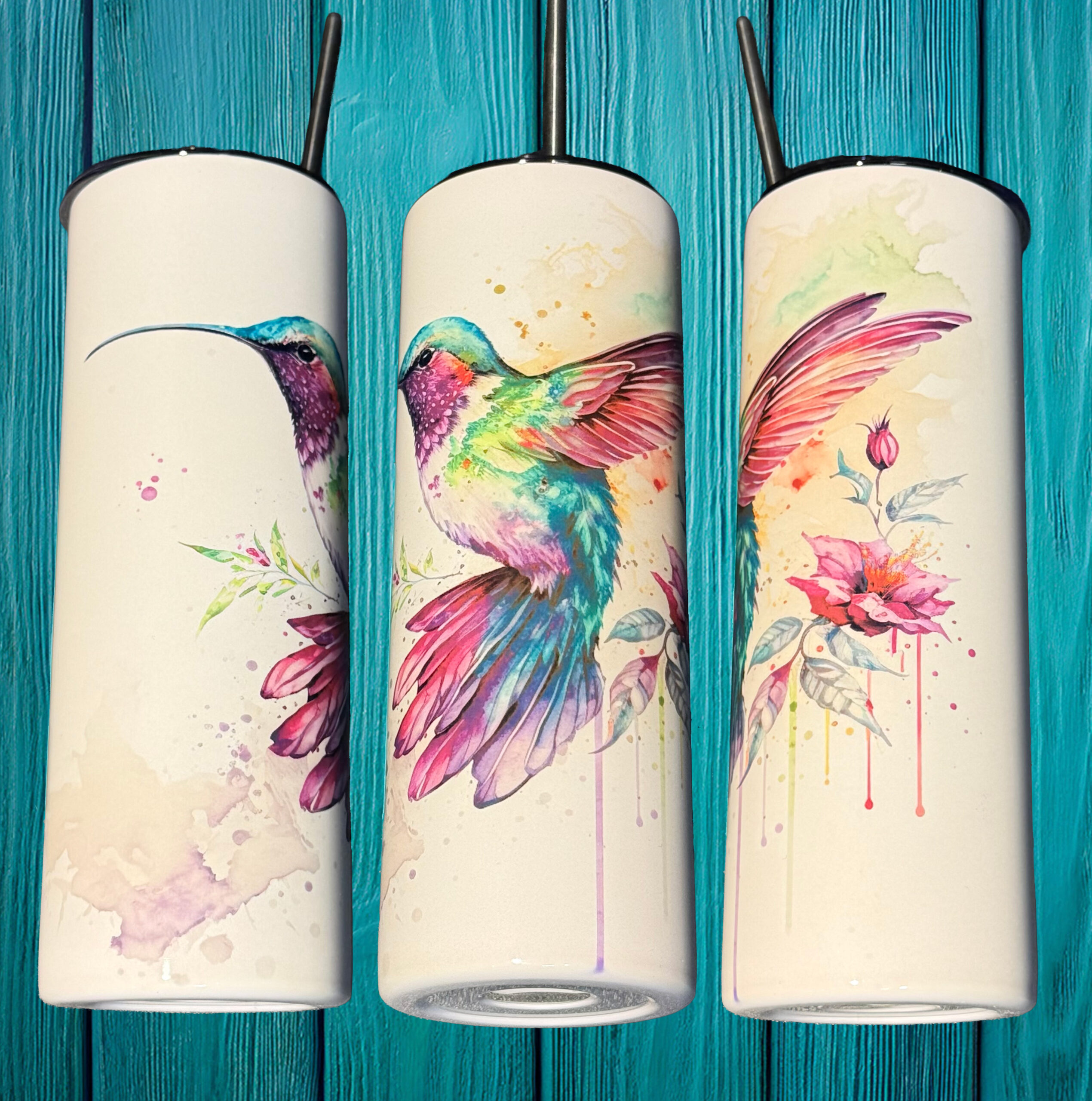 Three 20oz skinny tumblers featuring a vibrant watercolor hummingbird and floral design displayed against a turquoise wooden background.