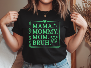 Woman wearing a black crew neck t-shirt with bright green graphic print reading Mama Mommy Mom Bruh in bold lettering inside a decorative bordered frame with a heart, sparkle stars, flowers, and smiley face accent icons
