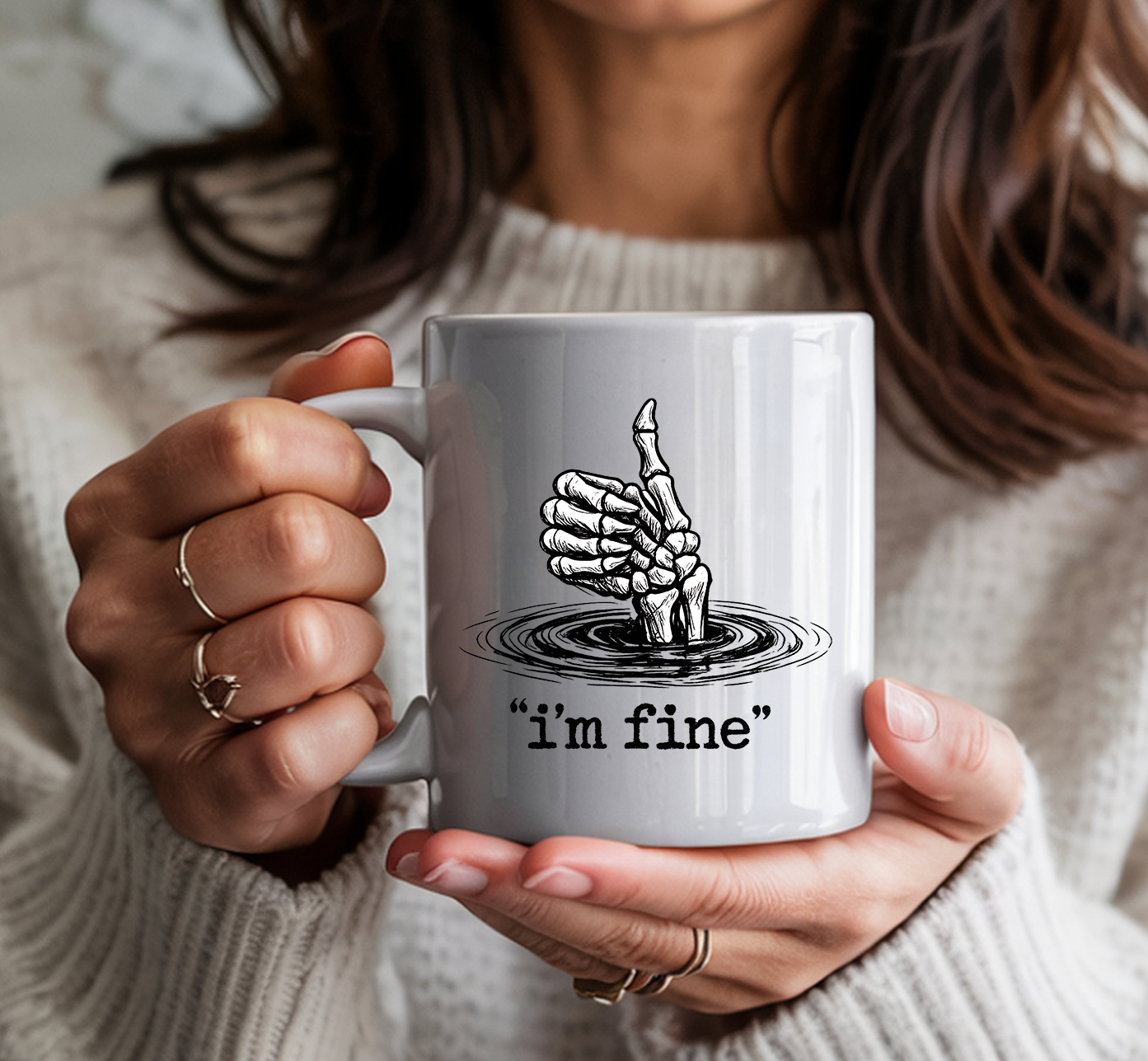 White ceramic coffee mug featuring a skeletal hand giving a thumbs up while sinking into water, with the text "i'm fine" in quotation marks — held by a person wearing a cozy cream sweater