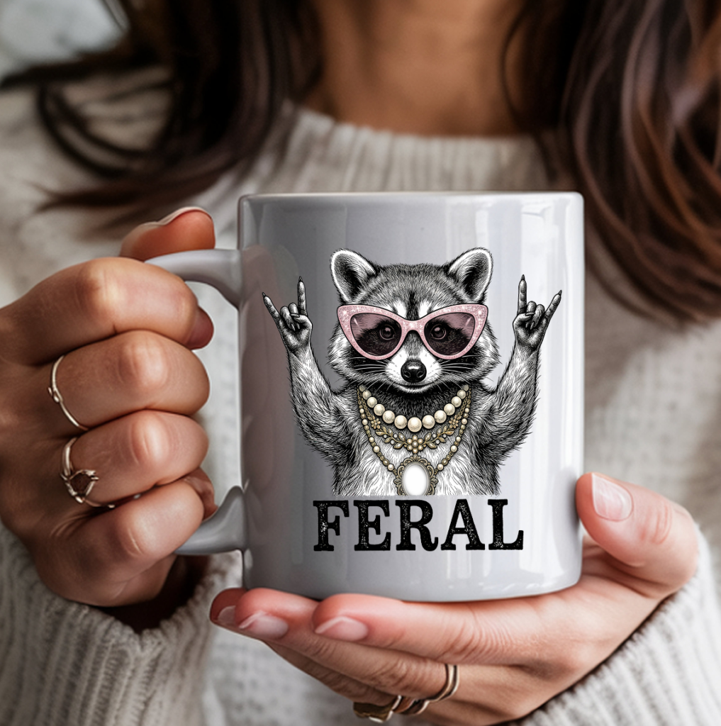 White ceramic coffee mug featuring a illustrated raccoon wearing pink cat-eye sunglasses and layered pearl and gold necklaces, throwing rock-on hand signs, with the word "FERAL" in bold text — held by a person in a cozy cream sweater