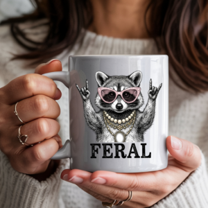 White ceramic coffee mug featuring a illustrated raccoon wearing pink cat-eye sunglasses and layered pearl and gold necklaces, throwing rock-on hand signs, with the word "FERAL" in bold text — held by a person in a cozy cream sweater