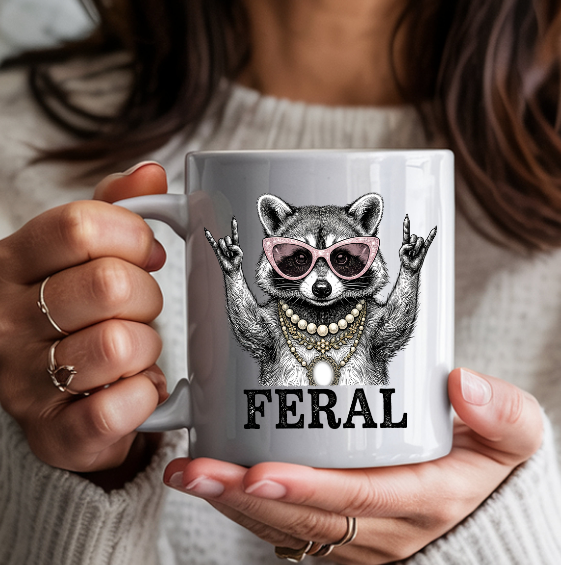 White ceramic coffee mug featuring a illustrated raccoon wearing pink cat-eye sunglasses and layered pearl and gold necklaces, throwing rock-on hand signs, with the word "FERAL" in bold text — held by a person in a cozy cream sweater