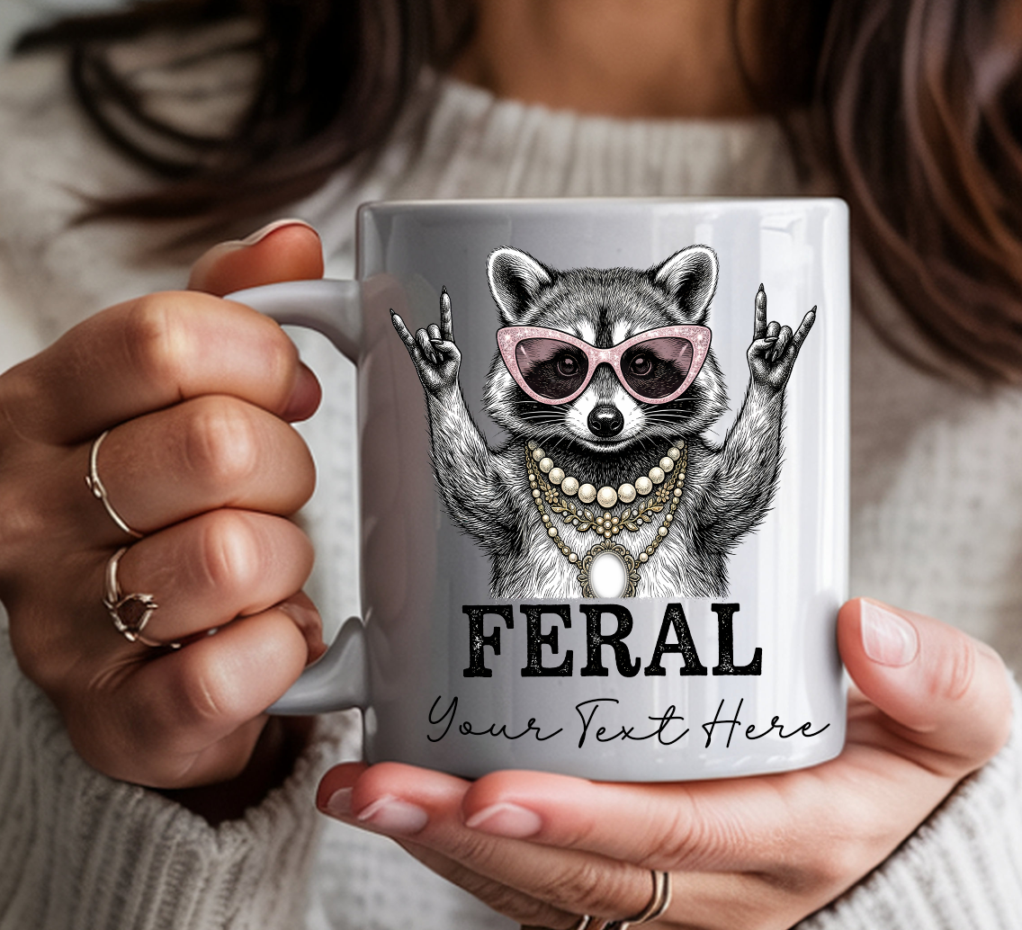 White ceramic coffee mug featuring a illustrated raccoon wearing pink cat-eye sunglasses and layered pearl and gold necklaces, throwing rock-on hand signs, with the word "FERAL" in bold text with personalization text — held by a person in a cozy cream sweater