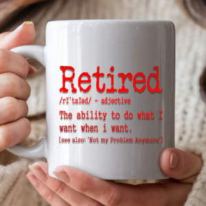 White coffee mug with sarcastic retirement definition: "Retired – The ability to do what I want when I want," in a red gradient color held in two hands against a cozy sweater background