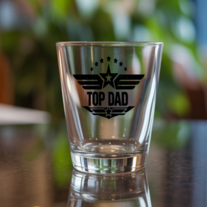 Top Dad shot glass with military star and wings graphic design on a dark reflective table with blurred green background