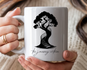 White ceramic coffee mug featuring a black silhouette illustration of a large tree with a spiral staircase winding up through its trunk, with the script text "the journey within" below. Held by a woman with a cozy sweater.