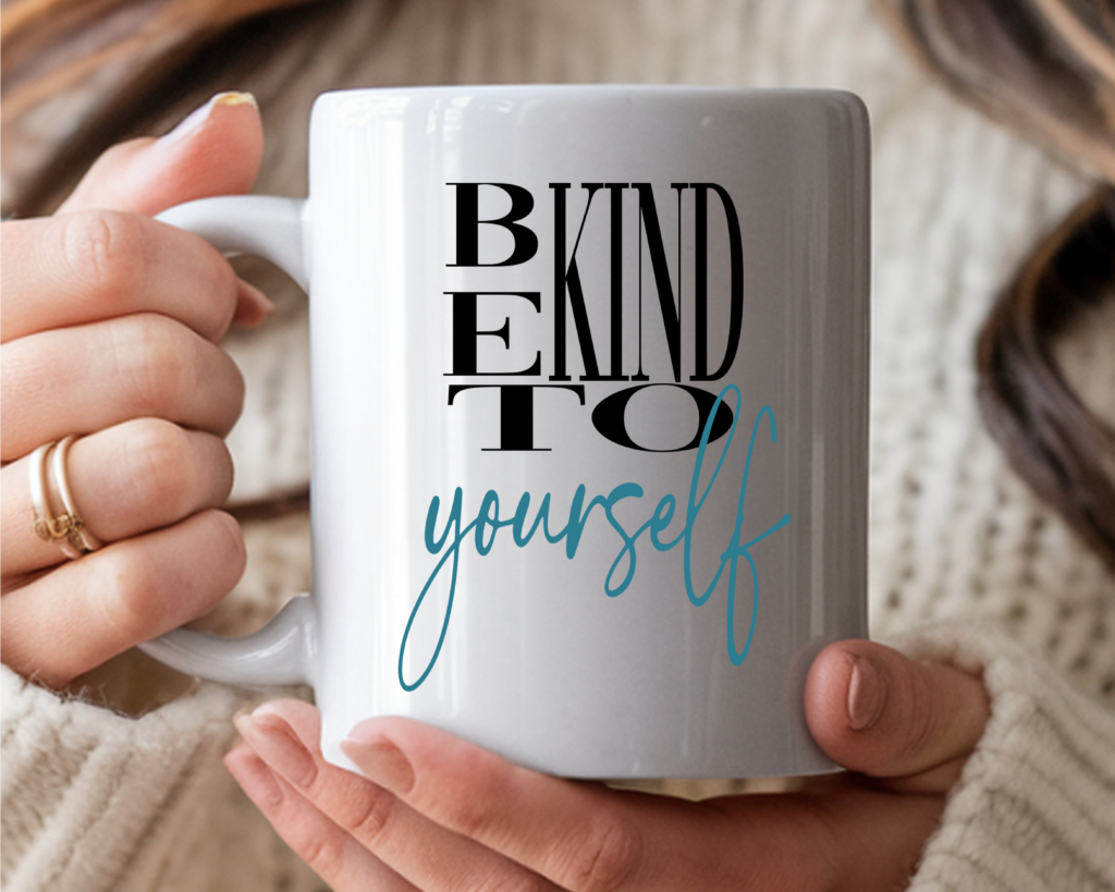 A woman's hands holding a white ceramic coffee mug printed with the bold black text "BE KIND TO" stacked vertically, and "yourself" written below in flowing teal script. She is wearing gold rings and a cream knit sweater, with a cozy warm background.