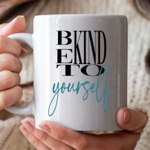 A woman's hands holding a white ceramic coffee mug printed with the bold black text "BE KIND TO" stacked vertically, and "yourself" written below in flowing teal script. She is wearing gold rings and a cream knit sweater, with a cozy warm background.
